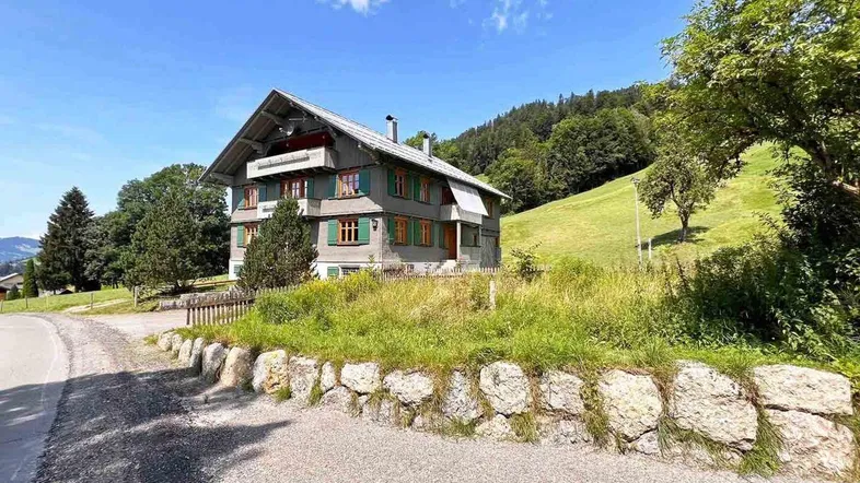 Traditionelles Haus im Alpenstil mit grünen Fensterläden und Bergblick, umgeben von grünen Wiesen.