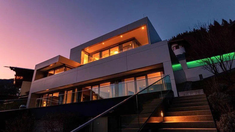 Moderne Villa mit beleuchteter Fassade und großen Glasfronten bei Sonnenuntergang in den Bergen.