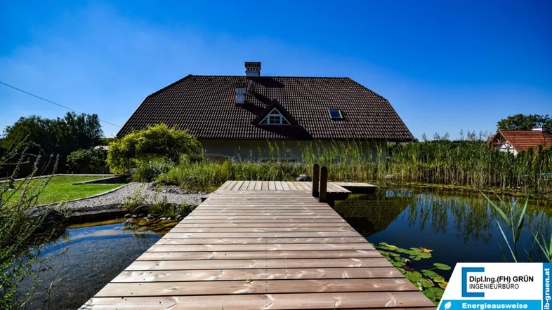 Haus mit idyllischem Teich und Holzsteg im Garten unter strahlend blauem Himmel.