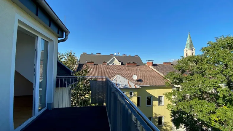 Geräumiger Balkon mit Blick über die Dächer der Stadt und einen Kirchturm bei strahlendem Sonnenschein.