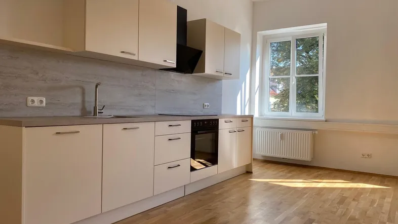 Helle Küche mit beigen Schränken, integriertem Backofen und Parkettboden, gut beleuchtet durch ein Fenster.