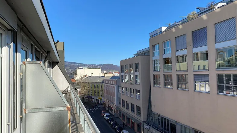 Blick vom Balkon auf eine belebte Straße mit Gebäuden und dahinterliegenden Hügeln unter blauem Himmel.