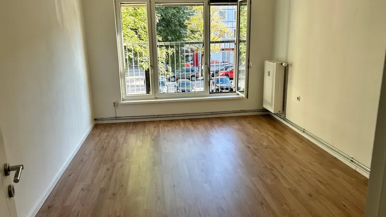 Helles Zimmer mit Laminatboden, weißen Wänden und Blick ins Grüne sowie auf die Straße.