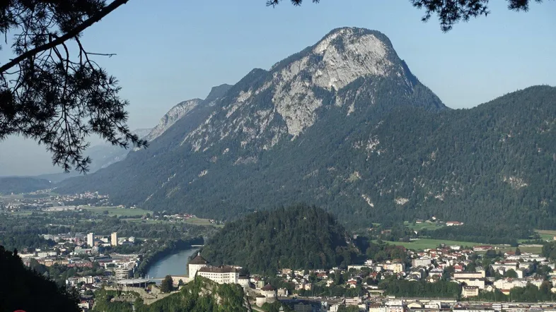 Panoramablick auf eine Stadt mit Fluss, Burg und majestätischen Bergen im Hintergrund unter klarem Himmel.