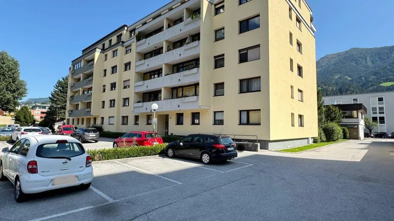 Mehrstöckiges Apartmentgebäude mit gelber Fassade und Parkplätzen vor einer Bergkulisse.