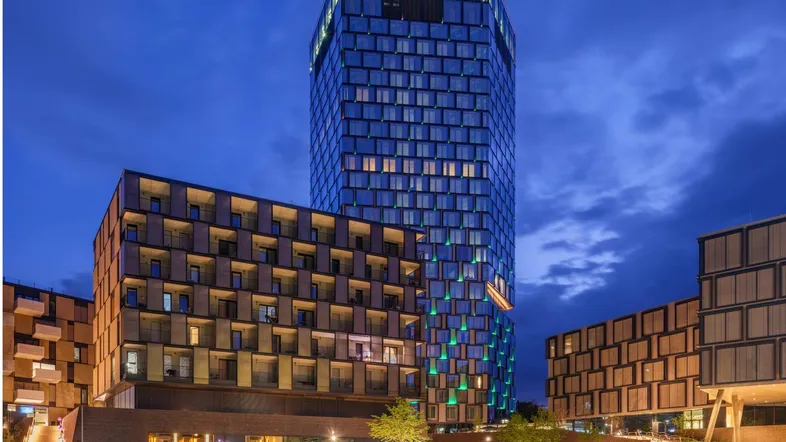 Modernes Hochhaus mit Glasfassade und beleuchteten Fenstern bei Nacht, umgeben von weiteren Gebäuden.