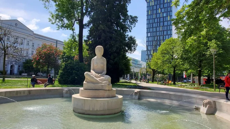 Ein belebter Stadtpark mit einem Brunnen und modernen Hochhäusern im Hintergrund unter blauem Himmel.