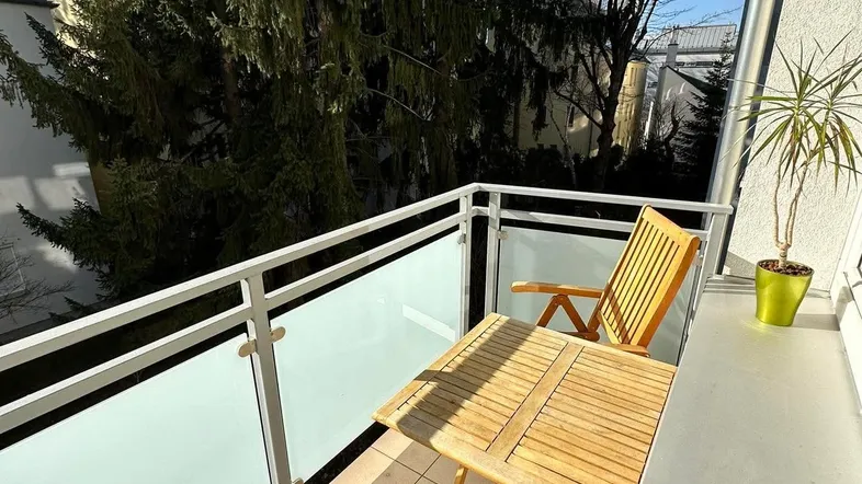 Sonniger Balkon mit Holztisch und Stühlen, umgeben von grünen Bäumen.