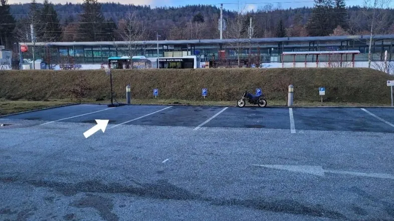 Großer Parkplatz mit Blick auf einen Bahnhof und bewaldete Hügel im Hintergrund.