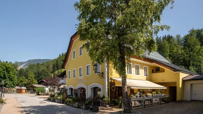 Gelbes Gebäude mit traditioneller Architektur, umgeben von Bäumen und Bergen, an einer Dorfstraße gelegen.