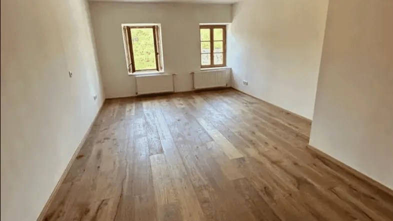 Helles, leeres Zimmer mit Holzboden und zwei Fenstern, bereit zur individuellen Gestaltung.