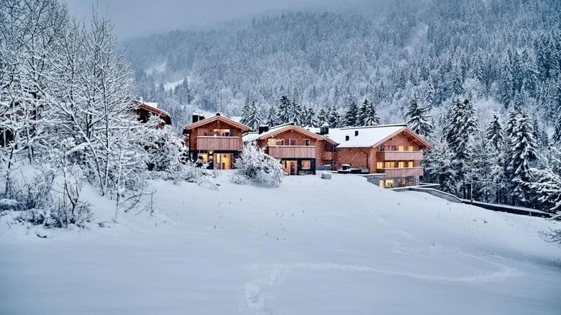 Moderne Chalets in einer verschneiten Berglandschaft, umgeben von Tannenbäumen und sanften Hügeln.