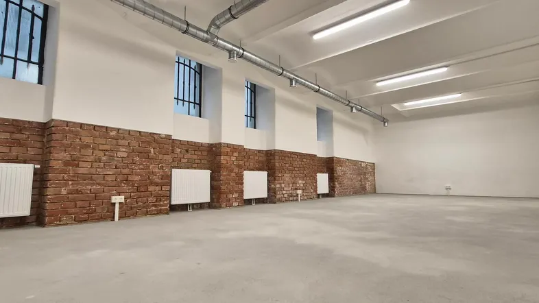 Großer, leerer Raum mit unverputzten Ziegelwänden, Betonboden und sichtbaren Lüftungsrohren an der Decke.