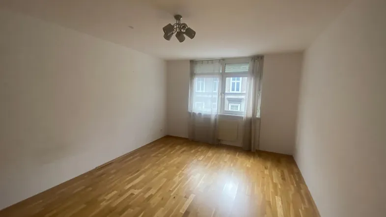 Helles, leeres Zimmer mit Parkettboden und einem Fenster, das Tageslicht hereinlässt.