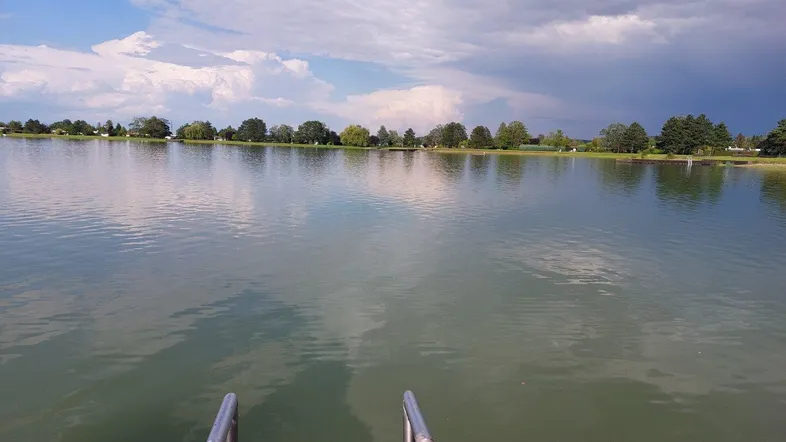 Blick auf einen ruhigen See mit Bäumen am Ufer unter einem bewölkten Himmel.