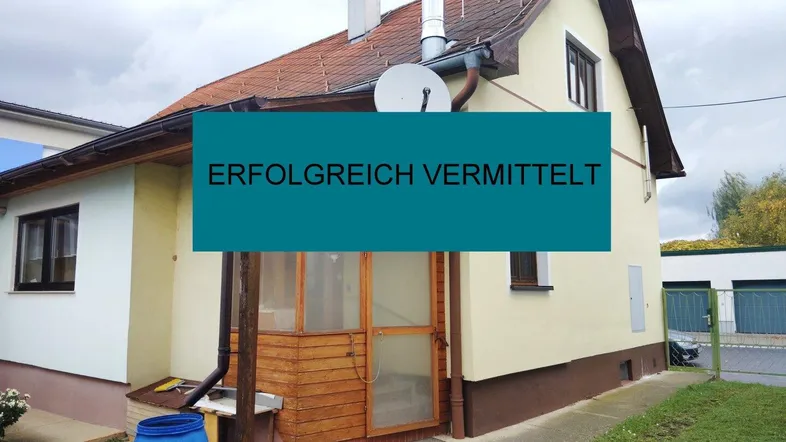 Kleines Einfamilienhaus mit Ziegeldach, heller Fassade und hölzernem Eingangsbereich, umgeben von Grünflächen.
