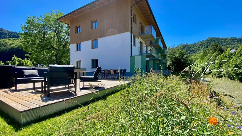 Moderne Wohnanlage mit Holzterrasse und Sitzgelegenheiten, umgeben von grüner Natur und Bergen.