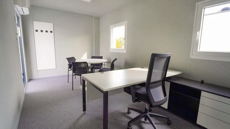 Modernes Büro mit Schreibtisch, Besprechungstisch und Teppichboden, gut beleuchtet durch Fenster.