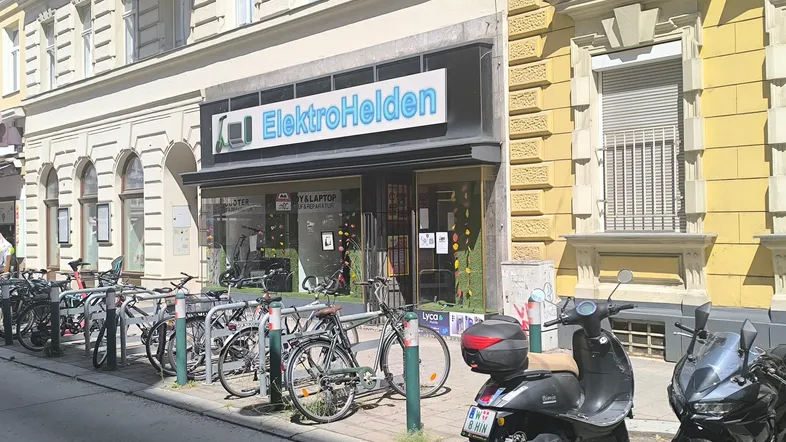 Fassade eines Geschäfts für Elektroartikel mit Fahrrädern und Rollern davor in einer belebten Straße.
