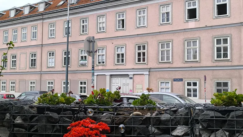 Mehrstöckiges Gebäude mit rosa Fassade und vielen Fenstern, umgeben von parkenden Autos und einer Gabionenmauer.