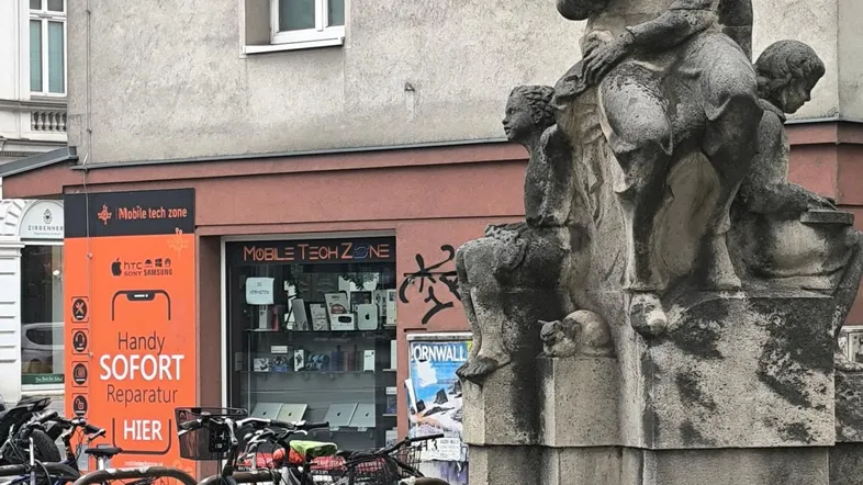 Blick auf eine belebte Straße mit einem historischen Denkmal, Fahrrädern und einem Gebäude mit Geschäften.