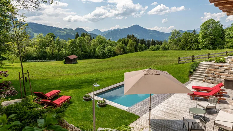 Großzügiger Außenbereich mit Pool, Holzterrasse und Liegestühlen, umgeben von grüner Natur und Bergblick.