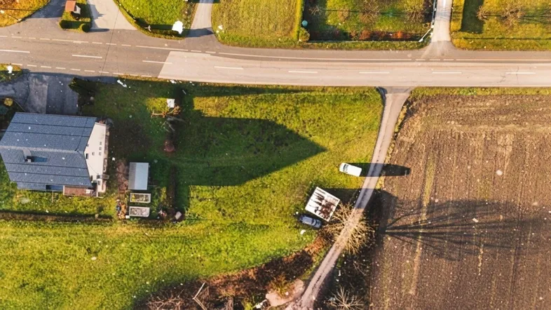 Luftaufnahme eines Hauses mit dunklem Dach, umgeben von grünen Wiesen und einem Acker, mit Straßen in der Nähe.
