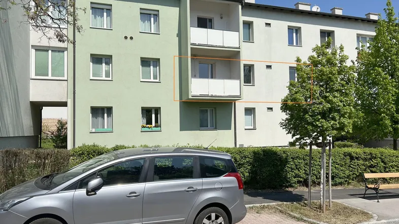 Mehrfamilienhaus mit grüner Fassade und Balkonen in sonniger Umgebung, ein Auto parkt davor.
