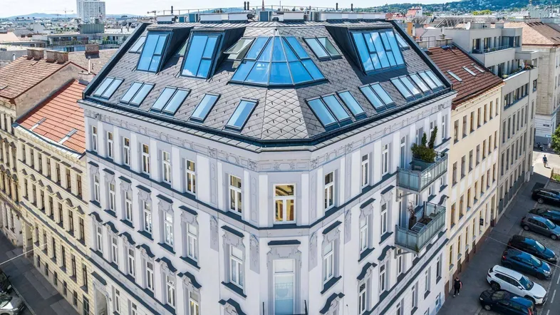 Luftaufnahme eines Eckgebäudes mit modernem Dachausbau und vielen Dachfenstern in urbaner Umgebung.