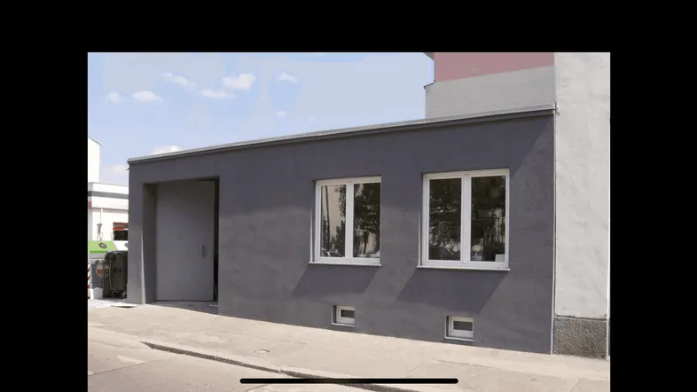 Graue Fassade eines funktionalen Gebäudes mit zwei Fenstern und einem großen Tor, direkt an einer Straße gelegen.