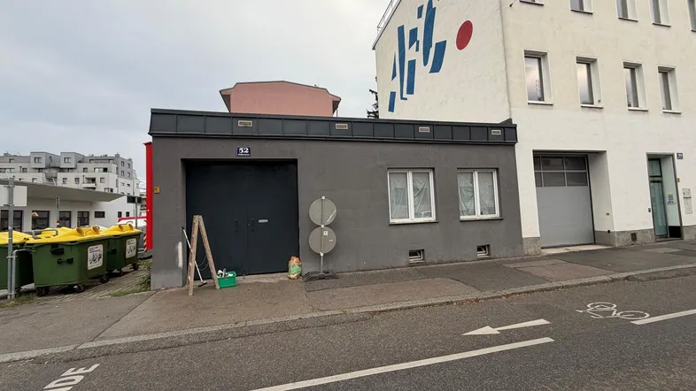 Graues Garagengebäude mit zwei Fenstern und einem großen Tor an einer Straße in städtischer Umgebung.