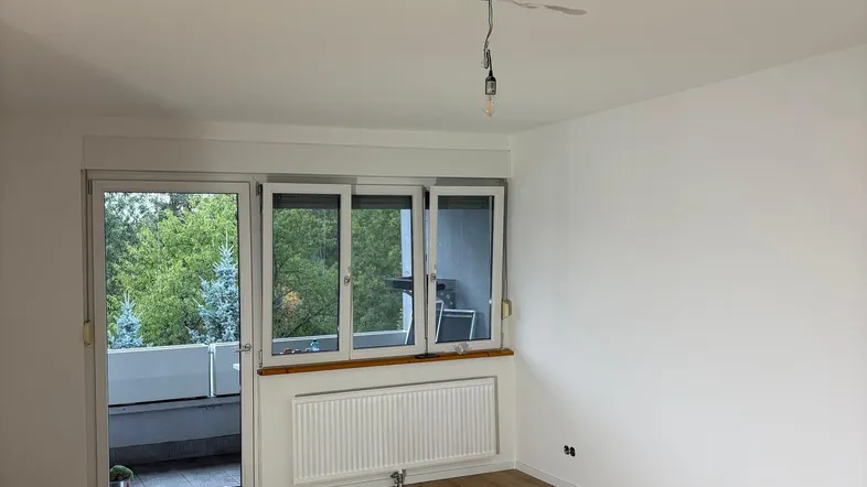 Helles, leeres Zimmer mit Laminatboden und direktem Zugang zu einem Balkon mit Blick ins Grüne.