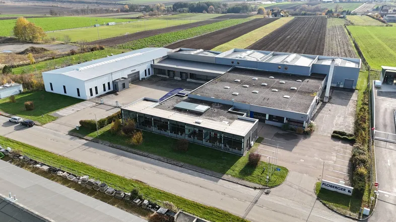 Luftaufnahme eines großen Gewerbekomplexes mit mehreren Hallen und umliegenden Feldern unter klarem Himmel.