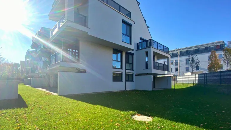 Modernes Mehrfamilienhaus mit weißer Fassade, großen Balkonen und gepflegter Rasenfläche bei Sonnenschein.