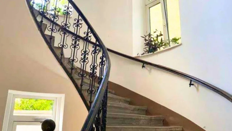 Elegantes Treppenhaus mit geschwungener Steintreppe und kunstvollem schmiedeeisernem Geländer, gut beleuchtet durch Fenster.