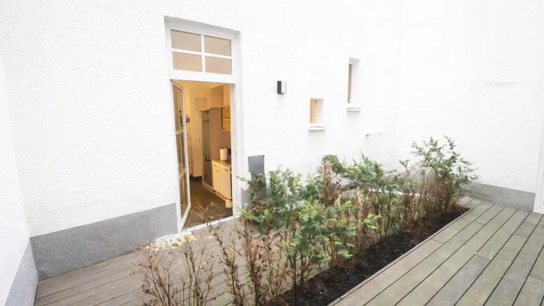 Kleine Terrasse mit Holzboden und Zugang zur Küche, umgeben von weißen Wänden und Pflanzen.