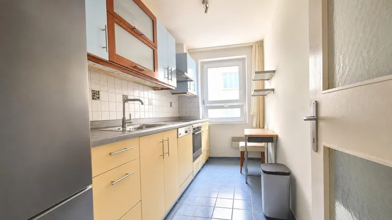 Schmale Küche mit blauen Fliesenboden, hellen Schränken und einem kleinen Esstisch am Fenster.