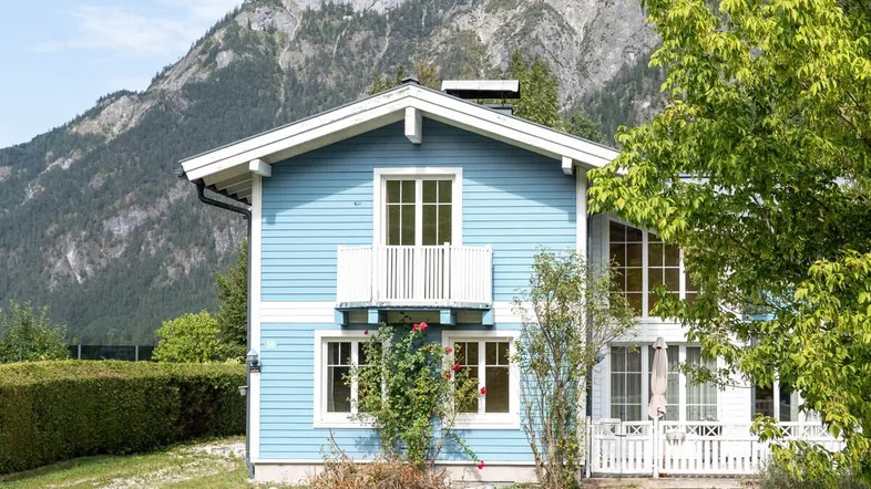 Blaues Haus mit weißem Balkon und Satteldach, umgeben von grüner Natur und Bergen.