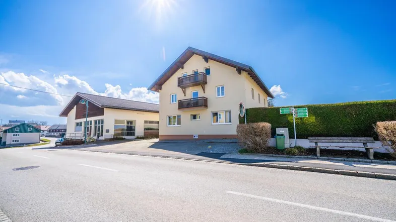 Mehrfamilienhaus mit Balkonen und angrenzendem Nebengebäude an einer sonnigen Straße.