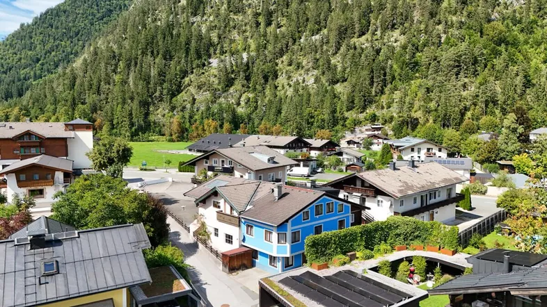 Dorfansicht mit Häusern, umgeben von grünen Bergen und Wäldern unter blauem Himmel.