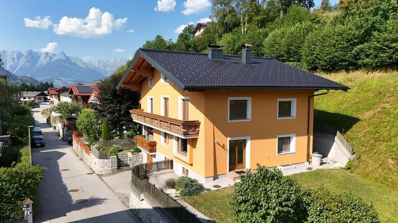 Gelbes Haus mit dunklem Dach und Holzbalkonen, umgeben von grüner Natur und Bergen.