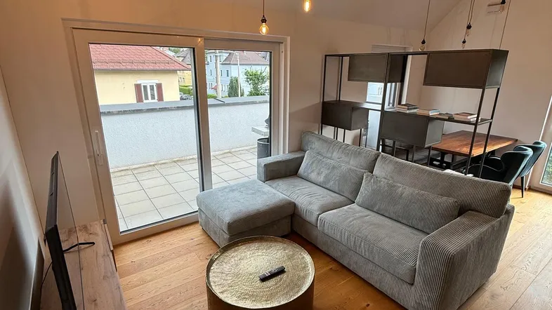 Modernes Wohnzimmer mit bequemer Couch, Holzboden und Zugang zu einer sonnigen Terrasse.