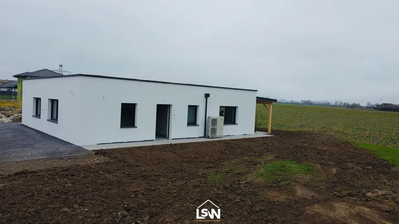 Weißes Einfamilienhaus mit Flachdach und Wärmepumpe, umgeben von unbebautem Land.