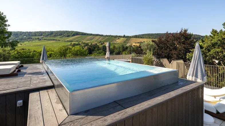 Moderner Infinity-Pool auf einer Holzterrasse mit weitem Blick über Weinberge und grüne Hügel an einem sonnigen Tag.
