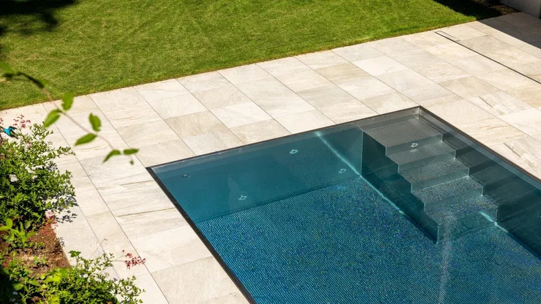 Moderner Swimmingpool mit gefliestem Bereich, umgeben von einer gepflegten Rasenfläche und Pflanzen.