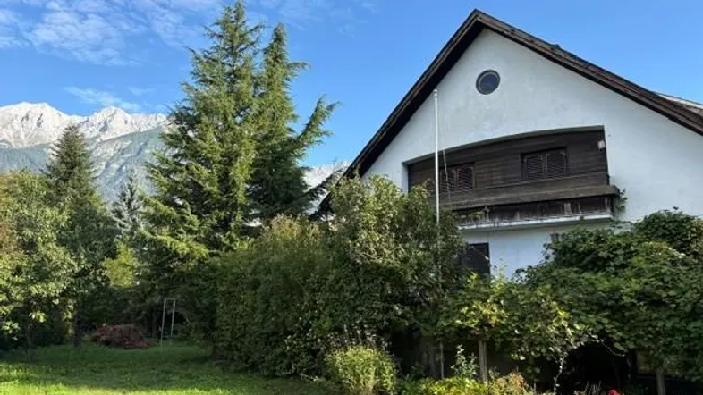 Traditionelles Haus mit Satteldach, umgeben von viel Grün und mit Bergblick auf die Alpen.