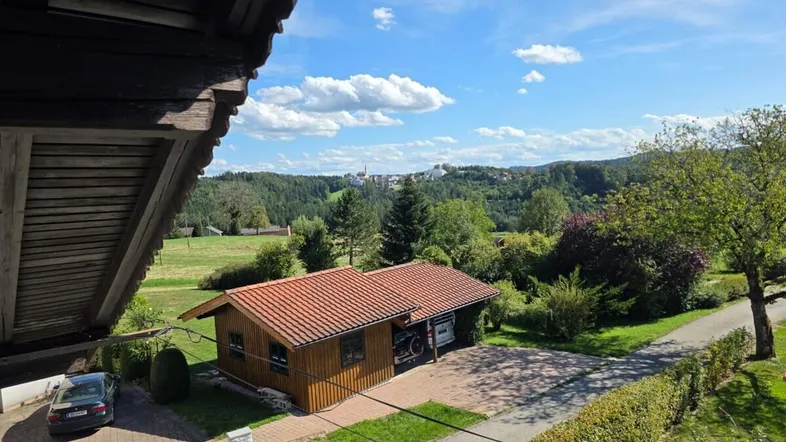 Panoramablick von einem Balkon auf ein Holzhaus, eine grüne Landschaft und ein Dorf in der Ferne unter blauem Himmel.