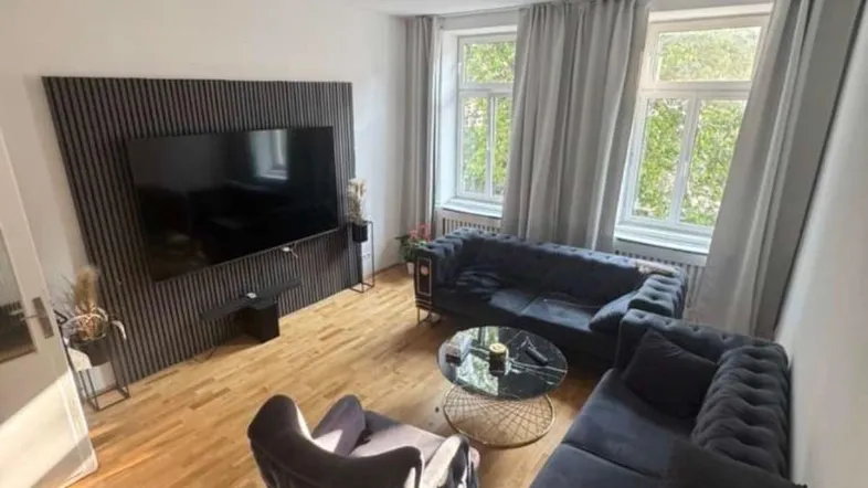 Helles Wohnzimmer mit dunklen Samtsofas, Holzboden und modernem TV-Bereich.