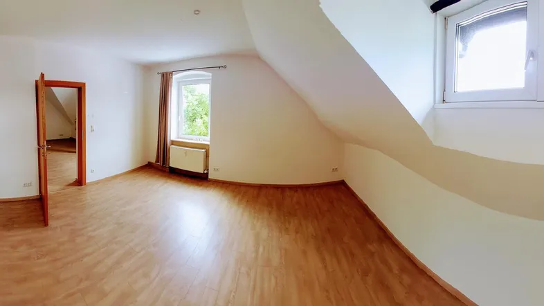 Heller Raum mit Dachschrägen, Laminatboden und natürlichem Lichteinfall durch große Fenster.