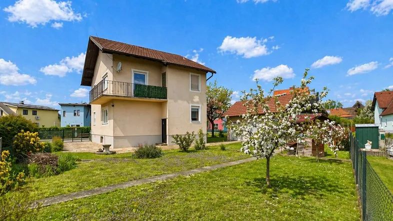 Ansicht des Hauses von der Gartenseite mit blühendem Baum und Rasenfläche unter blauem Himmel.